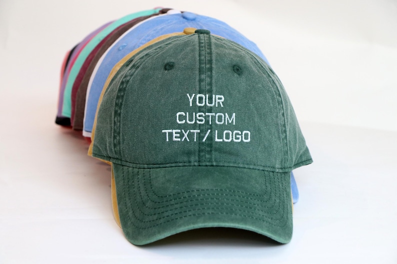 Peut inclure: Plusieurs casquettes de baseball de couleurs vari&eacute;es, dont vert, bleu et rose. La casquette avant est vert fonc&eacute; avec le texte "YOUR CUSTOM TEXT / LOGO" brod&eacute; en blanc. Les casquettes sont dispos&eacute;es en arc.