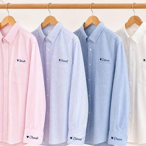 Pode incluir: Quatro camisas de botão de manga comprida, rosa, azul e branca, penduradas numa barra de madeira. Cada camisa tem uma palavra bordada no peito e no punho. As camisas são feitas de tecido listrado.