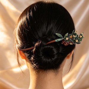 May include: A wooden hair stick featuring a decorative floral arrangement with dark green fabric leaves, small yellow flowers, and amber-coloured beads. The stem is wrapped in green ribbon, transitioning into a smooth, tapered brown wooden shaft.