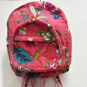 Puede incluir: Mochila acolchada rosa con un estampado floral en tonos azules, verdes y rosas. La mochila tiene un pequeño bolsillo frontal y correas ajustables con un diseño a rayas. La bolsa está hecha de un material acolchado suave.