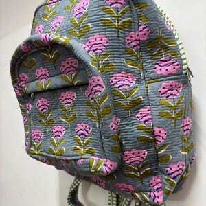 Puede incluir: Una mochila acolchada gris con un estampado floral. Las flores son rosas con tallos y hojas verdes. La mochila tiene un bolsillo frontal y correas a rayas. La cremallera es plateada.