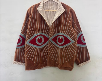 Puede incluir: Chaqueta marrón con diseños de ojos bordados en las mangas y el pecho. La chaqueta tiene cuello y puños a rayas. Los diseños de ojos son rojos con detalles azules y blancos. Cuelga de una percha de madera.