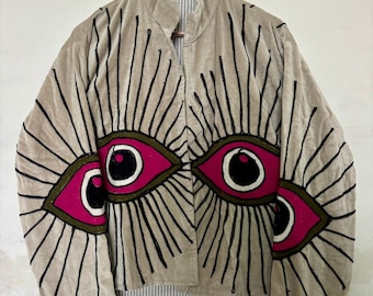 Puede incluir: Chaqueta de terciopelo beige con un diseño de ojos único. La chaqueta presenta tres ojos grandes con detalles rosas, negros y blancos, rodeados de líneas negras que irradian hacia afuera. La chaqueta está colgada en una percha de madera.