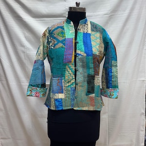 May include: A patchwork jacket in shades of teal, blue, and beige, with a mandarin collar and long sleeves. The jacket features a variety of patterned fabric squares sewn together, creating a unique and colorful design. The cuffs and collar are trimmed with a floral pattern.