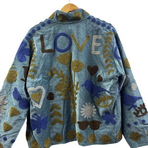 Peut inclure: Veste bleu clair avec broderies. Le mot "LOVE" est brodé au dos. La veste présente divers motifs brodés, notamment des fleurs, des oiseaux, des cœurs et des lunettes de soleil.