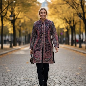 Op de afbeelding: Een vrouw draagt een lange jas met paisley-patroon, een zwarte broek en zwarte laarzen, lopend over een geplaveide straat. De jas heeft tinten roze, blauw en paars. De achtergrond toont bomen met gele bladeren.