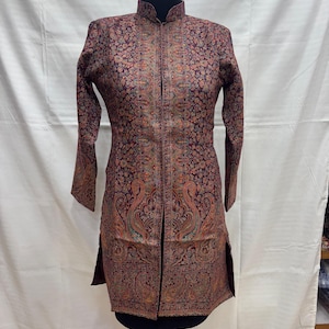Può includere: Una giacca lunga e fantasia nei toni del bordeaux, marrone e oro, esposta su un manichino. La giacca presenta un collo alto, maniche lunghe e un dettagliato design floreale e paisley. La giacca ha una cerniera frontale e spacchi laterali.