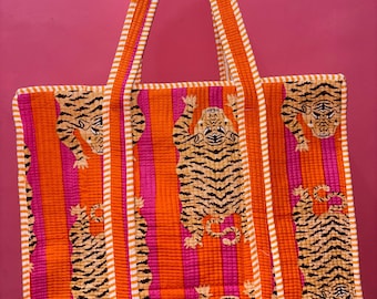 Könnte beinhalten: Eine gesteppte Tragetasche mit Tigerdruck. Die Tasche hat ein sich wiederholendes Muster von Tigern vor einem Hintergrund aus vertikalen orangefarbenen und rosa Streifen. Die Griffe und die Zierleisten sind orange mit weißen Streifen.