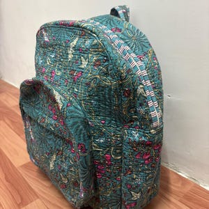 Puede incluir: Mochila acolchada en color turquesa con un estampado floral en rosa y dorado. La mochila tiene un asa superior, un bolsillo frontal y cierre de cremallera. La tela parece ser de algodón.