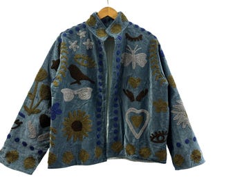 May include: A blue velvet jacket with embroidered floral, bird, and eye designs. The jacket features a collared neckline and long sleeves. The embroidery includes shades of brown, gold, and white, creating a unique, artistic look.