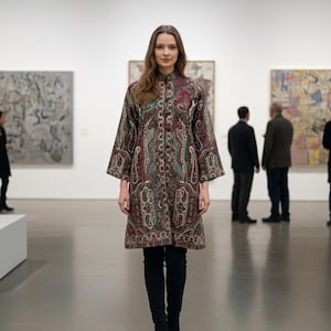 Peut inclure: Un long manteau boutonné avec un motif cachemire dans les tons de marron, rouge et vert. Le manteau a de larges manches et est porté avec un pantalon et des bottes noirs. La photo est prise dans une galerie d'art.
