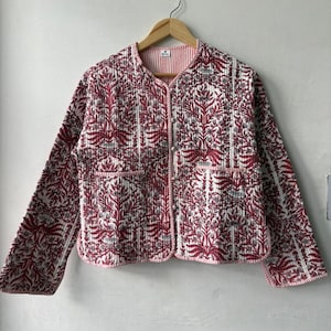 Puede incluir: Chaqueta acolchada con estampado floral en tonos rojos y blancos. La chaqueta tiene cuello redondo, mangas largas y dos bolsillos delanteros. La chaqueta está colgada de una percha de madera.