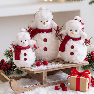 May include: Three white plush snowman figurines with orange carrot noses, black eyes, and red scarves. Each snowman wears a white fuzzy hat and has two brown buttons on the front. They are positioned on a wooden sled with red berry accents.