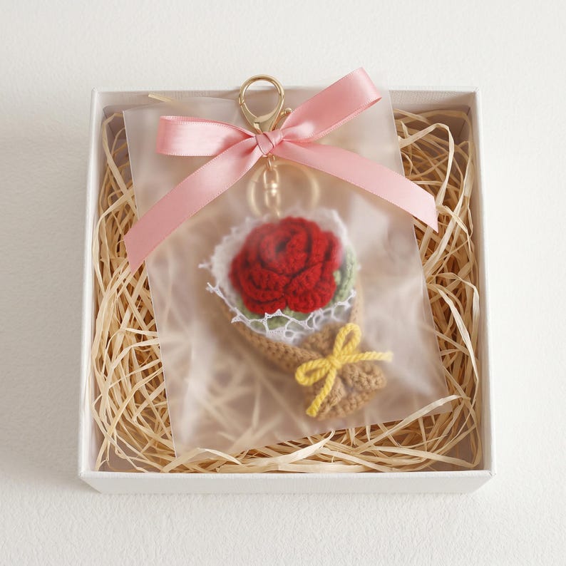 Puede incluir: Un llavero de ramo de rosas de ganchillo en una caja de regalo. La rosa roja est&aacute; rodeada de hojas verdes y encaje blanco, envuelta en un cono marr&oacute;n y atada con un lazo amarillo. El llavero tiene un cierre dorado y una cinta rosa.