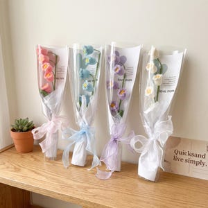Puede incluir: Cuatro ramos individuales de flores de lirio de los valles tejidas a ganchillo en rosa, azul, morado y blanco. Cada tallo está envuelto en papel blanco con una cinta transparente y lleva el texto love more impreso en el envoltorio.
