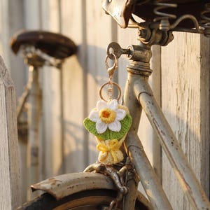 Puede incluir: Un llavero de ganchillo hecho a mano con un ramo de flores blancas y amarillas con hojas verdes, atado con una cinta amarilla. El llavero est&aacute; sujeto a un cierre dorado y cuelga de una bicicleta vintage.