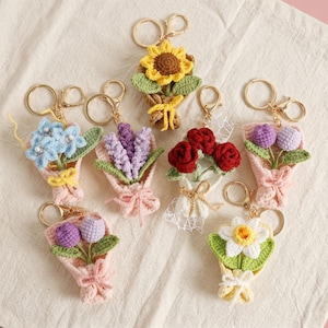 Puede incluir: Una colecci&oacute;n de llaveros de ramo de flores hechos a mano con ganchillo. Cada llavero presenta un dise&ntilde;o de flor diferente, incluyendo girasoles, rosas y lavanda, en varios colores. Los ramos est&aacute;n envueltos en papel rosa o blanco y atados con una cinta, sujetos a un llavero dorado.