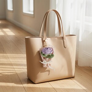 Puede incluir: Un bolso tote beige con asas a juego y un cierre dorado. Un peque&ntilde;o charm de ramo de flores de ganchillo en tonos morados, verdes y rosas est&aacute; sujeto a la bolsa. La bolsa est&aacute; sobre un suelo de madera.