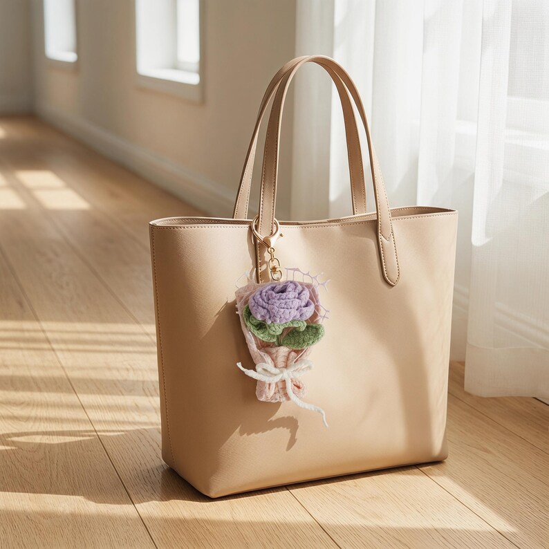 Puede incluir: Un bolso tote beige con asas a juego y un cierre dorado. Un peque&ntilde;o charm de ramo de flores de ganchillo en tonos morados, verdes y rosas est&aacute; sujeto a la bolsa. La bolsa est&aacute; sobre un suelo de madera.