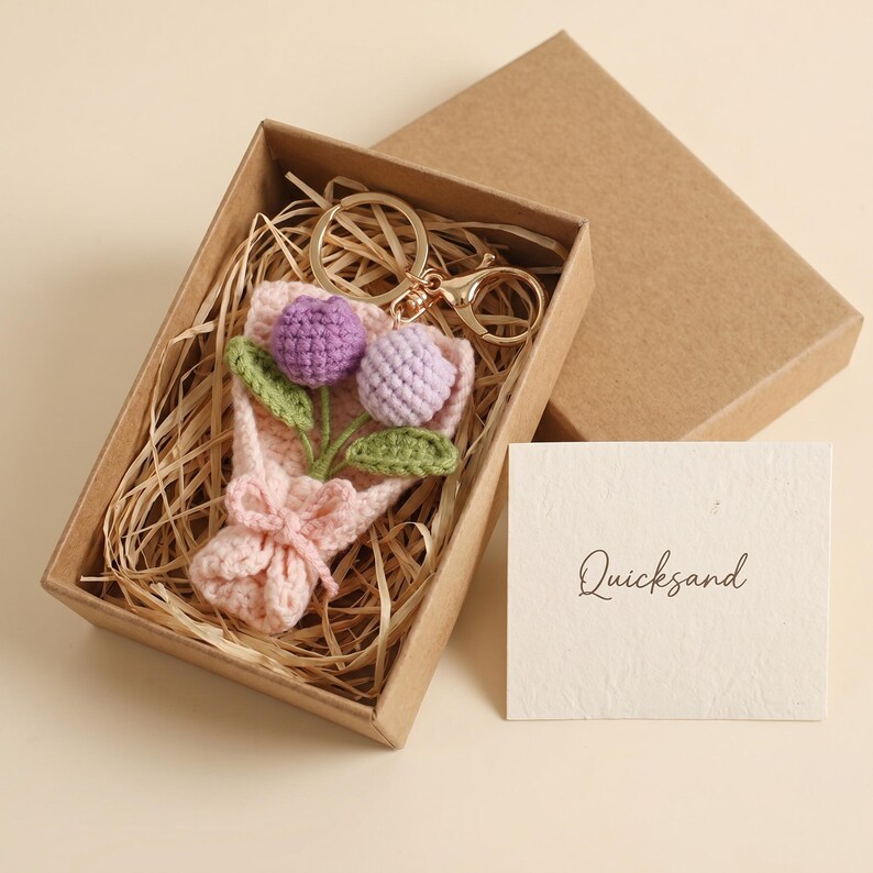 Puede incluir: Un llavero de ramo de flores de ganchillo en una caja de regalo. El ramo presenta flores moradas y lavanda con hojas verdes, atadas con un lazo rosa. El llavero es dorado. La caja es marr&oacute;n y contiene paja.