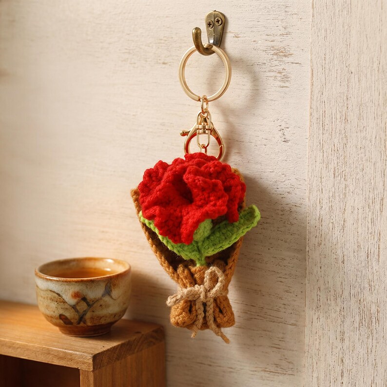 Puede incluir: Un llavero de ganchillo hecho a mano con un ramo de flores rojas con hojas verdes, envuelto en un material marr&oacute;n similar al papel y atado con una cuerda. El llavero est&aacute; sujeto a un anillo y un cierre de metal dorado.