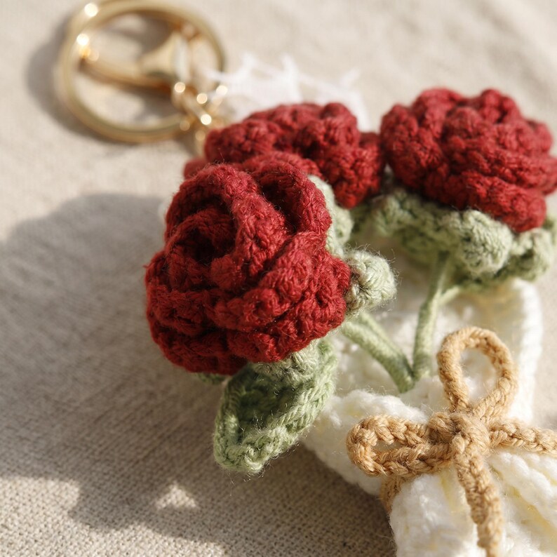 Puede incluir: Un llavero hecho a mano con un ramo de rosas rojas de ganchillo con hojas y tallos verdes. Las rosas est&aacute;n unidas a una base blanca de ganchillo, atada con un lazo marr&oacute;n. Un llavero dorado est&aacute; unido en la parte superior.