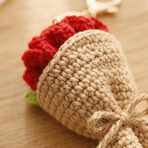 Puede incluir: Un llavero de ramo de ganchillo hecho a mano con una flor roja vibrante y hojas verdes, envuelto en un cono beige texturizado. El ramo est&aacute; atado con un lazo de cuerda natural, creando un accesorio encantador.