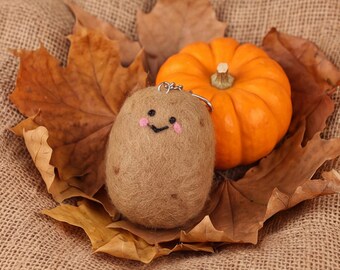 May include: A tan needle-felted potato keychain with a stitched black smile and pink cheeks, featuring a silver metal keyring attached to the top, positioned next to a small orange pumpkin and several dried brown maple leaves.