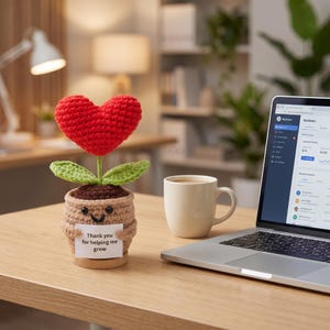 Puede incluir: Una planta de ganchillo con una flor roja en forma de corazón, hojas verdes y una maceta color canela con una cara sonriente. La maceta sostiene un letrero blanco que dice "Thank you for helping me grow". El objeto está sobre un escritorio.