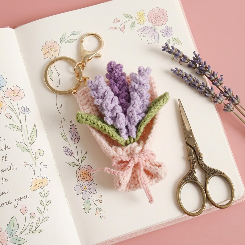 Puede incluir: Un llavero de ganchillo hecho a mano con un ramo de flores de lavanda en tonos morados y verdes, atado con un lazo rosa. El llavero tiene un cierre dorado. La imagen tambi&eacute;n incluye unas tijeras peque&ntilde;as y una ramita de lavanda.