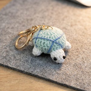May include: A crochet turtle keychain featuring a pale mint green shell with blue yarn detailing, a white head, white flippers, and small black bead eyes. The turtle is attached to a gold-toned metal split ring and a lobster claw clasp.
