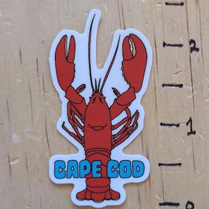 May include: A red lobster sticker with the words "CAPE COD" in blue letters. The lobster has a smiling face and is approximately 2 inches tall.