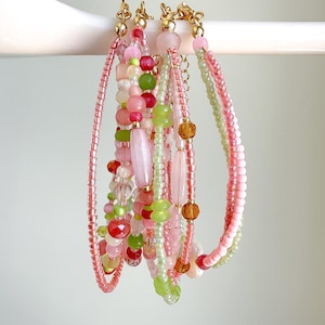May include: A collection of beaded bracelets in shades of pink, green, and gold. The bracelets feature various bead shapes and sizes, including round, oval, and faceted beads. The bracelets are hanging from a white bar.