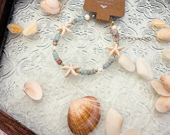 Op de afbeelding: Een handgemaakte armband met zeesterbedels, blauwe, gouden en witte kralen. De armband wordt gepresenteerd met schelpen op een getextureerd glazen oppervlak. Een klein label leest "Handmade with love."