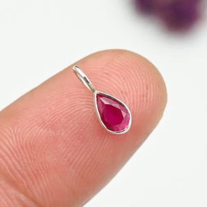 Könnte beinhalten: Ein kleiner, tropfenförmiger Anhänger mit einem leuchtend roten Edelstein, der in einem silberfarbenen Metall gefasst ist. Der Edelstein ist facettiert und fängt das Licht ein. Der Anhänger ist als Halskette konzipiert.