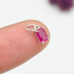 May include: A small, rectangular, ruby-colored gemstone pendant with a silver setting. The pendant is held by a silver loop. The gemstone is a vibrant pink-red color. The pendant is shown against a blurred background.