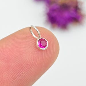 Könnte beinhalten: Ein kleiner Silberanhänger mit einem runden, leuchtend rosa Edelstein. Der Anhänger wird von einem Finger gehalten, mit einem verschwommenen Hintergrund aus lila Blumen. Der Edelstein ist in einer Lünettenfassung gefasst, und der Anhänger hat eine kleine Schlaufe für eine Kette.
