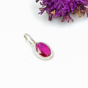 Puede incluir: Un colgante de plata con una vibrante gema de rubí facetada de forma ovalada. El rubí está engastado en un bisel de plata pulida, y el colgante tiene un lazo simple para una cadena. Un racimo de flores moradas está en el fondo.