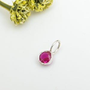 Op de afbeelding: Een zilveren hanger met een ronde, helder roze edelsteen. De hanger heeft een lus voor een ketting. De edelsteen is gezet in een zilveren bezel. Een gele bloem staat op de achtergrond.