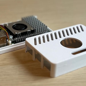 May include: A white computer case with a slotted top and a circular opening, next to a circuit board with a fan and heat sink. The case is designed to house electronic components. The image is taken on a wooden surface.