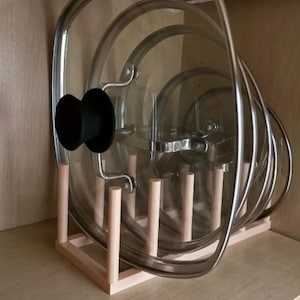 May include: A wooden pot lid organizer with multiple clear glass lids with metal rims and black handles. The organizer has a rectangular base and vertical wooden pegs to hold the lids in place. The lids are stored inside a wooden cabinet.