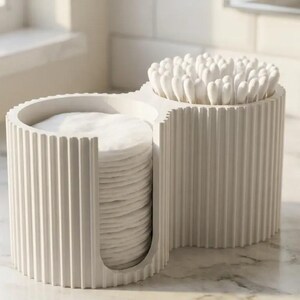May include: A white, ribbed, dual-compartment bathroom organiser. One side features a U-shaped cutout holding a stack of round white cotton pads, while the other side is a cylindrical container filled with white cotton swabs.