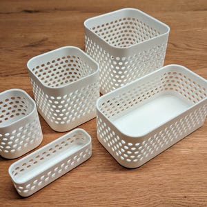 May include: A set of five white plastic storage baskets in varying shapes and sizes, each featuring a perforated grid pattern on the sides. The collection includes two square containers, one rectangular bin, one small square cup, and one long narrow tray.