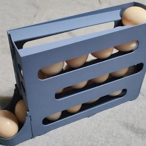 May include: A slate blue plastic egg dispenser rack featuring a gravity-fed design with multiple horizontal slots. The structure holds numerous light beige chicken eggs, with one egg resting in the front dispensing tray at the base of the unit.