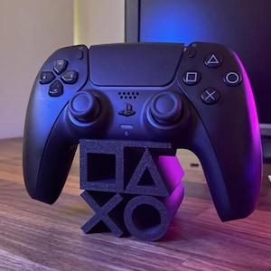 May include: A black PlayStation 5 controller resting on a black stand with the PlayStation symbols. The controller has a symmetrical design with buttons and joysticks. The stand is made of a foam-like material.