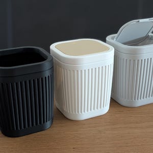 May include: Three small square waste bins with vertical ribbed textures on the sides. One bin is black and open, one is white with a tan lid, and one is light grey with an open hinged lid. Each container features rounded corners and a compact design.