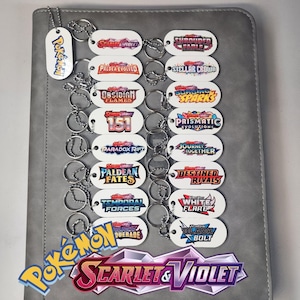 Könnte beinhalten: Eine Sammlung weißer, ovaler Schlüsselanhänger mit verschiedenen Pokémon-Designs und Logos. Die Schlüsselanhänger sind an silbernen Ketten befestigt und auf einer grauen Oberfläche angeordnet. Das Bild enthält das Pokémon Scarlet & Violet Logo.
