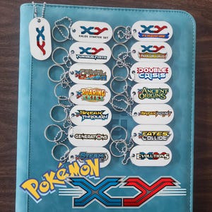 Puede incluir: Una colección de llaveros blancos con forma de chapa de perro con varios logotipos y textos de la serie Pokémon XY. Los llaveros muestran logotipos como "Kalos Starter Set", "Furious Fists", "Flashfire", "Phantom Forces", "Double Crisis", "Ancient Origins", "Break Through", "Breakpoint", "Generations", "Steam E", "Fates Collide" y "Evolutions".