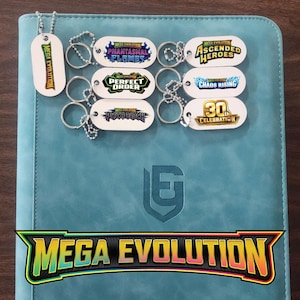 May include: A collection of white oval keychains with metal ball chains, each featuring colorful logos including Mega Evolution, Phantasmal Flames, Ascended Heroes, Perfect Order, Chaos Rising, Pitch Black, and 30th Celebration, resting on a teal binder.