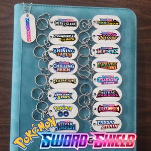 May include: A collection of white, oval-shaped dog tags with various Pokémon Sword & Shield series names printed on them, including "Rebel Clash," "Darkness Ablaze," and "Champion's Path." Each tag is attached to a silver ball chain. The tags are displayed on a teal background.